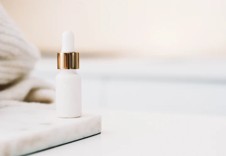 A white dropper bottle with a gold collar on white marble